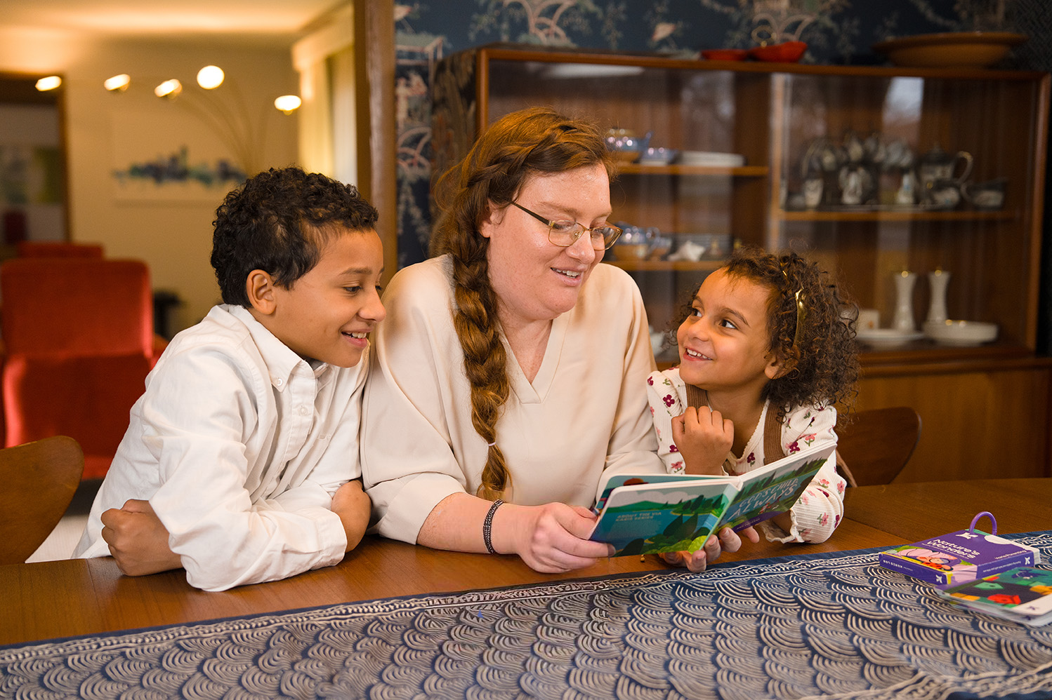 Woman reading God's Child Always by Karis Parenting to two children at a table in a room with shelves in the background.